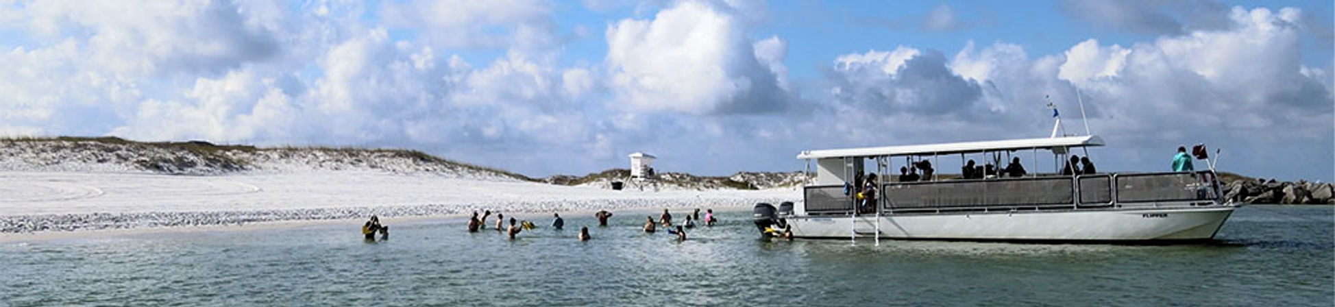 Panoramic white-sand beach with dunes, swimmers wading in shallow turquoise water and a covered tour boat anchored near shore under a bright partly cloudy sky.
