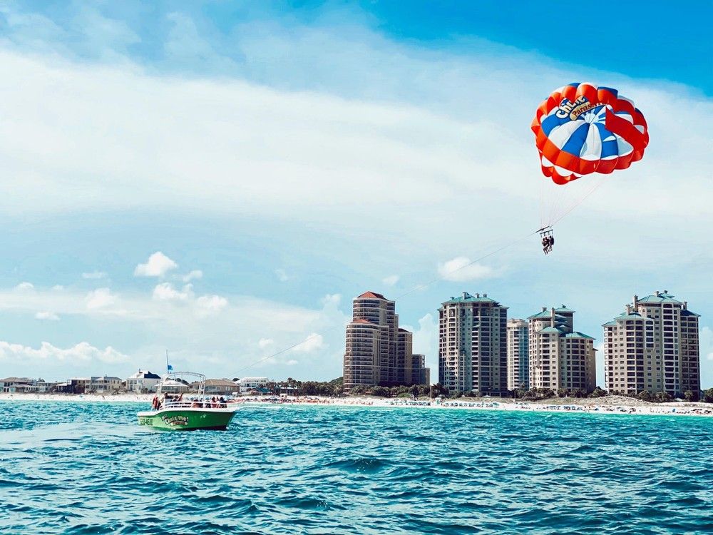 Colorful parasail with two riders soaring over turquoise ocean, towed by a green motorboat off a sandy beach lined with oceanfront condo towers under a bright blue sky.