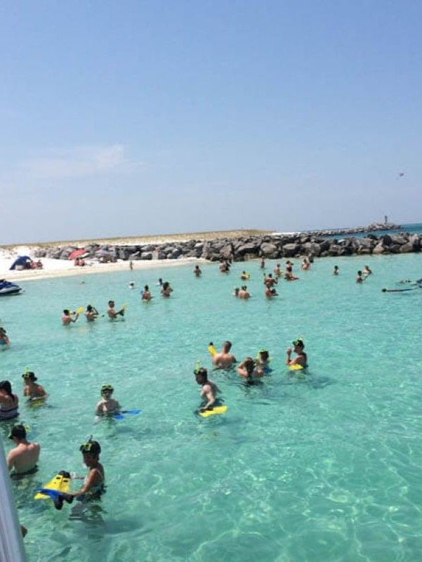 Crowded tropical beach lagoon with clear turquoise water, swimmers and snorkelers in masks and fins, sandy shore with umbrellas and a rocky jetty under a bright blue sky