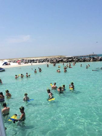 Crowded tropical beach lagoon with clear turquoise water, swimmers and snorkelers in masks and fins, sandy shore with umbrellas and a rocky jetty under a bright blue sky