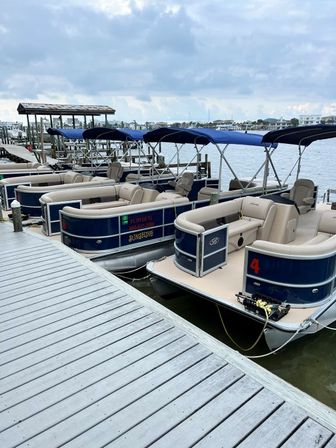 Line of navy-and-beige pontoon boats with blue canopies moored at a wooden marina dock, calm harbor waters and waterfront homes under a cloudy sky