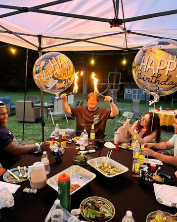 Outdoor backyard birthday party at dusk under a white canopy, friends seated around a table with takeout trays, salads and drinks, string lights and two large clear Happy Birthday balloons, central guest holding two lit fire torches for a playful celebration.