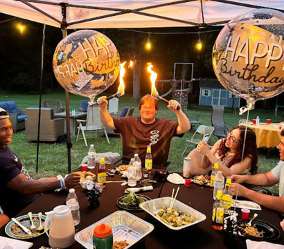 Outdoor backyard birthday party under a canopy at dusk, friends gathered around a table of plates and drinks, two large "Happy Birthday" balloons, and a celebrant holding two flaming torches while guests laugh and take photos.
