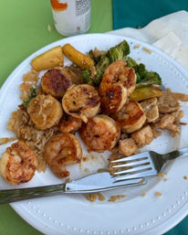 Sizzling grilled shrimp and seared scallops over fried rice with broccoli and zucchini on a white plate at an outdoor picnic table, fork and knife visible