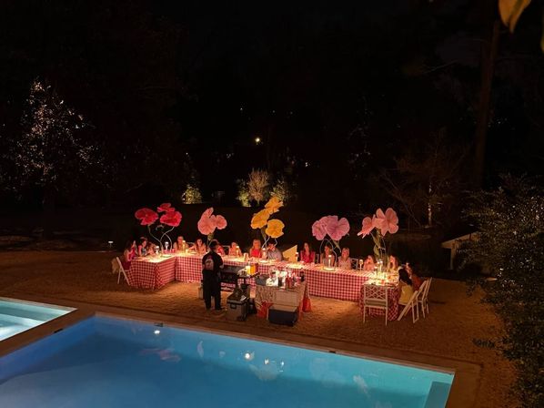 Nighttime outdoor poolside dinner in a garden with a long red-gingham banquet table, guests seated under oversized pink and yellow paper flower decorations, candlelit lanterns and a server at a food cart, blue pool reflecting warm ambient light.