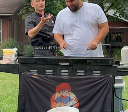 Backyard barbecue cookout with a chef in a black jacket gesturing while a man in a white t‑shirt and playful chef hat flips food on a flat‑top grill under trees, banner with cartoon chef on the grill front