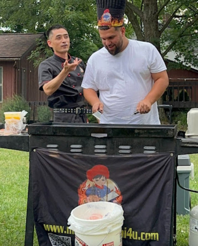 Backyard barbecue cookout with a chef in a black jacket gesturing while a man in a white t‑shirt and playful chef hat flips food on a flat‑top grill under trees, banner with cartoon chef on the grill front