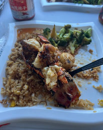 Grilled lobster tail atop egg fried rice with sautéed broccoli in a white takeout-style plate — charred, juicy seafood dish.