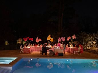 Poolside backyard nighttime dinner party with long red-gingham tables, oversized illuminated paper flowers, guests dining under warm lights and their reflections in the swimming pool