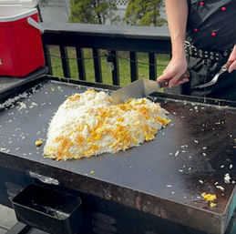 Backyard flat-top griddle on a wooden deck with a pile of rice and scrambled eggs being chopped and mixed with a metal spatula by a cook.