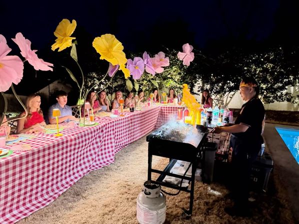 Evening backyard poolside party with a long red-gingham table, oversized colorful paper flowers above, guests seated and a chef grilling with visible flames.