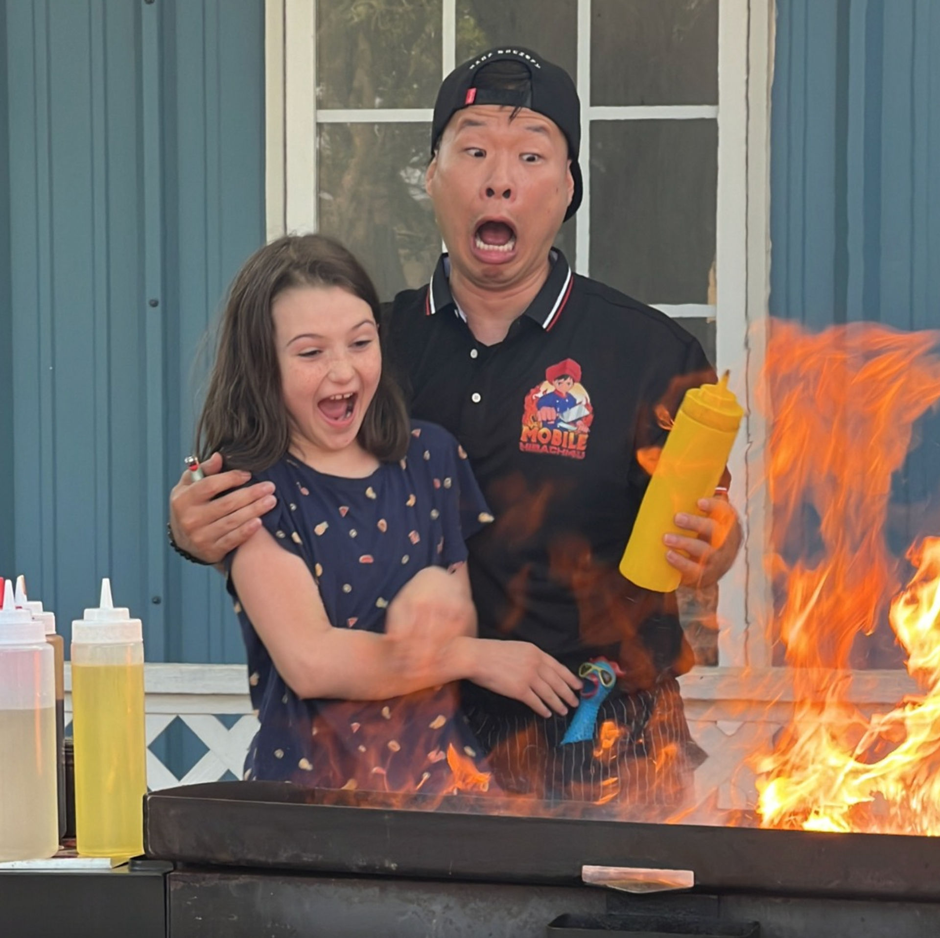 Surprised chef and young girl at an outdoor cooking demonstration, large orange flames erupting from a flat-top grill as the chef holds a yellow squeeze bottle, condiment bottles and a blue building with a window behind them.