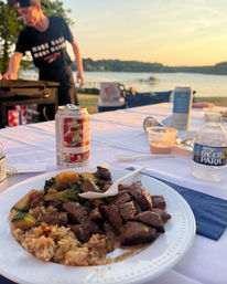 Lakeside summer cookout: paper plate of grilled steak bites, rice and vegetables with a plastic fork, canned drink and bottled water on a picnic table, person at a grill and a dock at sunset