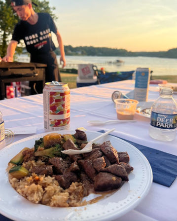 Lakefront sunset picnic: paper plate of grilled steak bites with seasoned rice and sautéed zucchini and broccoli, plastic fork, canned drink and water bottle on a table, blurred cook at a grill and dock across the lake in the background.