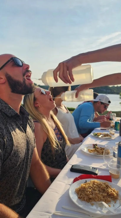 Playful group at a lakeside summer picnic leaning back at a long table as someone pours drinks from squeeze bottles into open mouths; plates of food, drink cans and a lake in the background.