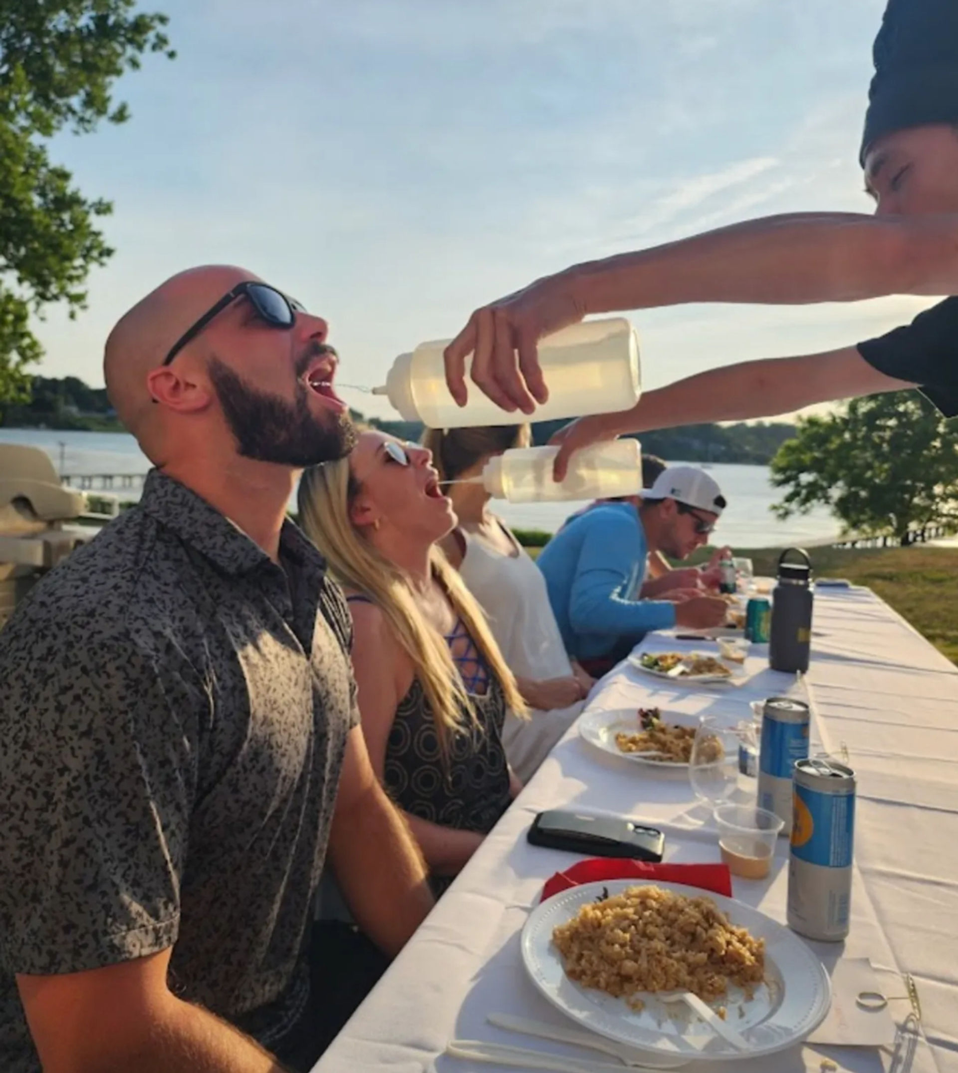 Playful group at a lakeside summer picnic leaning back at a long table as someone pours drinks from squeeze bottles into open mouths; plates of food, drink cans and a lake in the background.