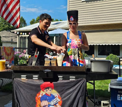 Two people at a portable hibachi flat-top in a suburban backyard under an American flag — one person cooking while a friend in a colorful sundress and tall novelty hat sips a drink at a summer outdoor BBQ.