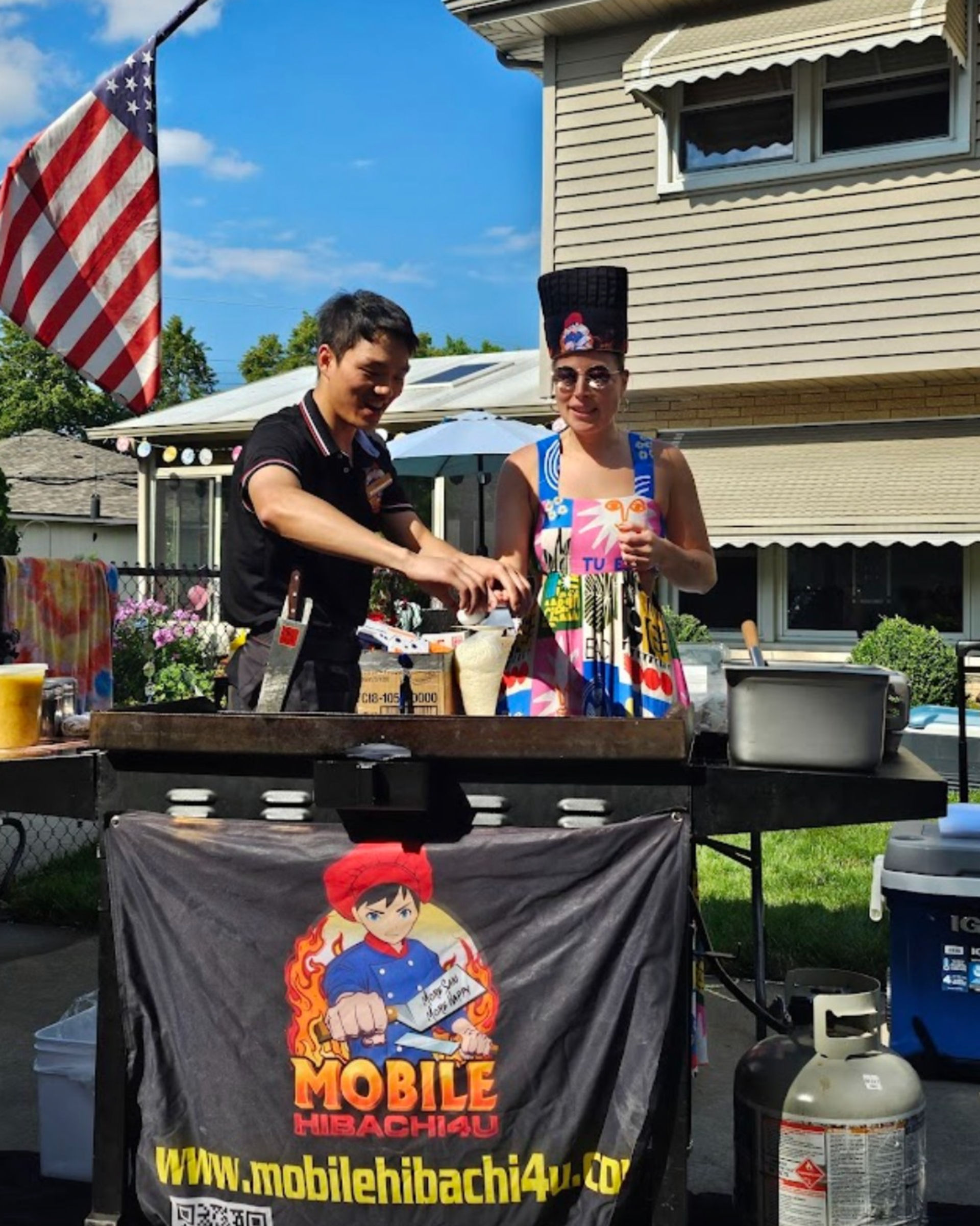 Two people at a portable hibachi flat-top in a suburban backyard under an American flag — one person cooking while a friend in a colorful sundress and tall novelty hat sips a drink at a summer outdoor BBQ.