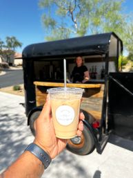 Iced latte in a clear plastic cup with a straw held up to the camera in front of a black mobile coffee trailer on a sunny suburban street.