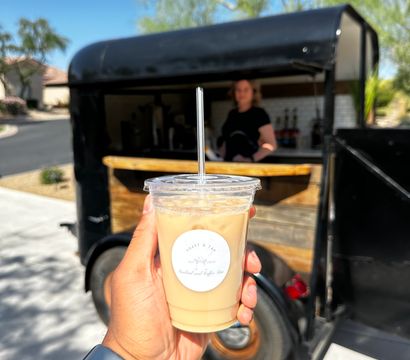 Iced latte in a clear plastic cup with a straw held up to the camera in front of a black mobile coffee trailer on a sunny suburban street.