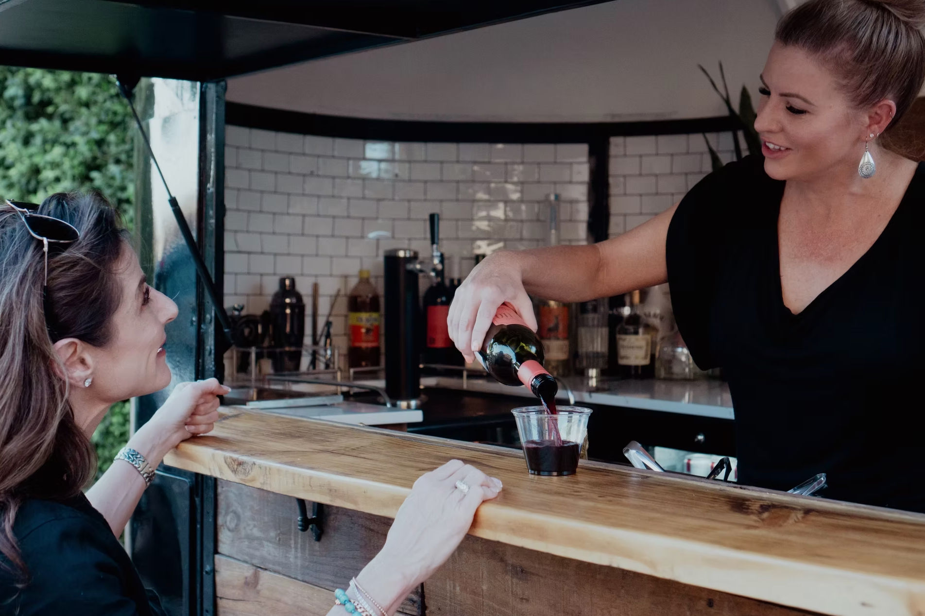 Bartender pouring red wine into a plastic cup for a smiling customer at an outdoor mobile bar with a wooden counter and white tile backsplash.
