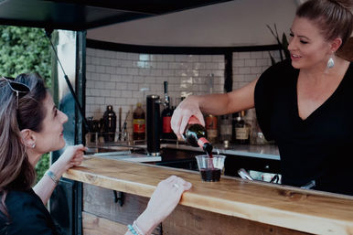 Bartender pouring red wine into a plastic cup for a smiling customer at an outdoor mobile bar with a wooden counter and white tile backsplash.