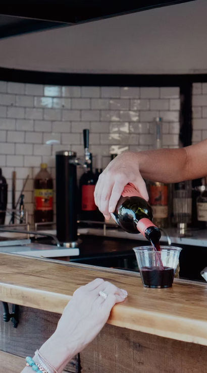 Bartender pouring red wine into a plastic cup for a smiling customer at an outdoor mobile bar with a wooden counter and white tile backsplash.