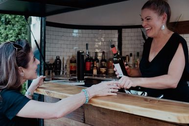 Smiling bartender hands a bottle of red wine to a customer across a wooden pop-up bar counter, with a tiled back wall and bottles visible in the background.