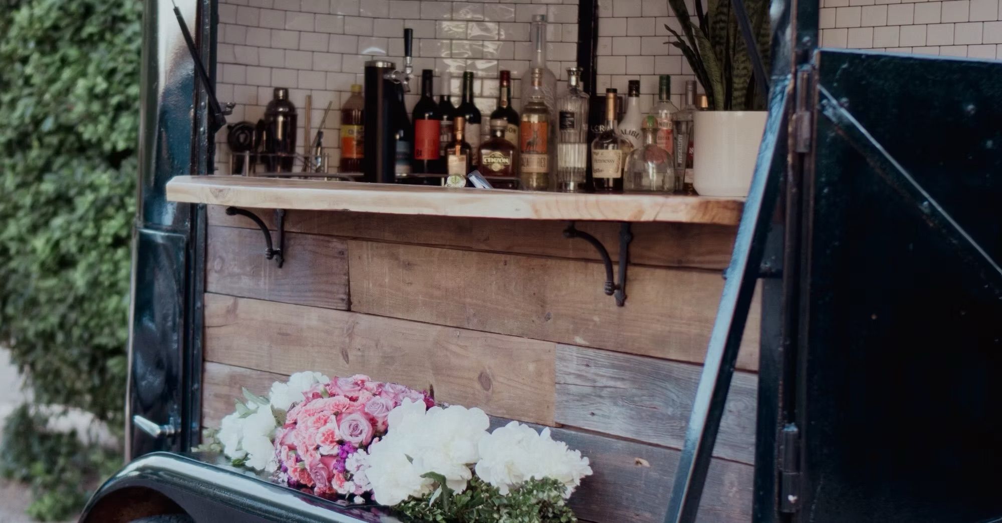 Charming outdoor vintage mobile bar trailer with wooden counter and subway-tile backsplash, bottles on the shelf, a potted plant and pink and white flower bouquets.