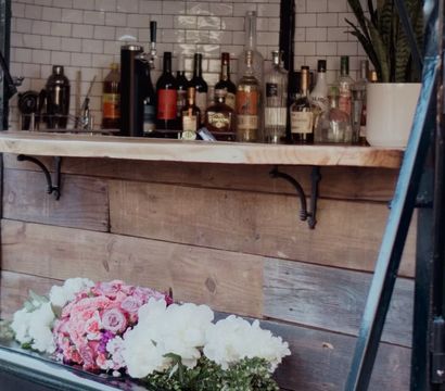 Charming outdoor vintage mobile bar trailer with wooden counter and subway-tile backsplash, bottles on the shelf, a potted plant and pink and white flower bouquets.