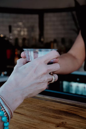 Close-up of hands exchanging a clear plastic cup of dark drink over a wooden bar counter; foreground wrist wearing turquoise beaded bracelets and a gold ring.
