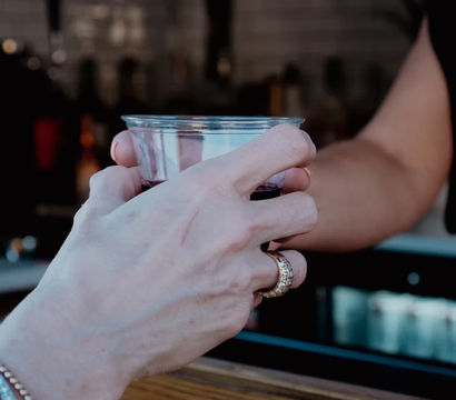 Close-up of hands exchanging a clear plastic cup of dark drink over a wooden bar counter; foreground wrist wearing turquoise beaded bracelets and a gold ring.