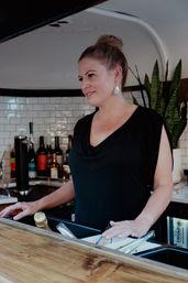 Woman bartender with hair in a bun leaning at a wooden bar counter inside a modern cafe with white subway tile, wine bottles and a potted plant