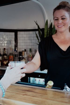 Smiling bartender hands a plastic cup cocktail to a customer across a wooden bar counter with bottles and a white subway-tile backsplash