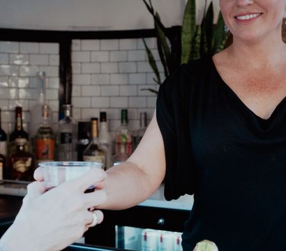Smiling bartender hands a plastic cup cocktail to a customer across a wooden bar counter with bottles and a white subway-tile backsplash