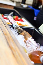 Sparkling rosé bottle chilled in crushed ice beside trays of strawberries and orange slices on a wooden bar