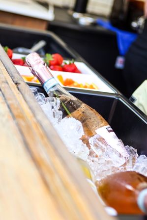 Sparkling rosé bottle chilled in crushed ice beside trays of strawberries and orange slices on a wooden bar