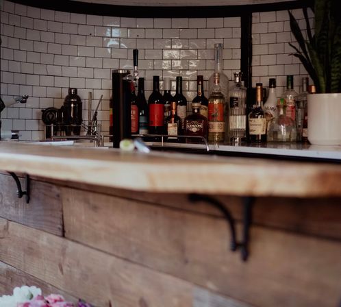 Rustic wooden bar counter with glossy white subway-tile backsplash, chrome beer tap, assorted liquor bottles, cocktail shaker and a small potted plant — cozy indoor bar setup.