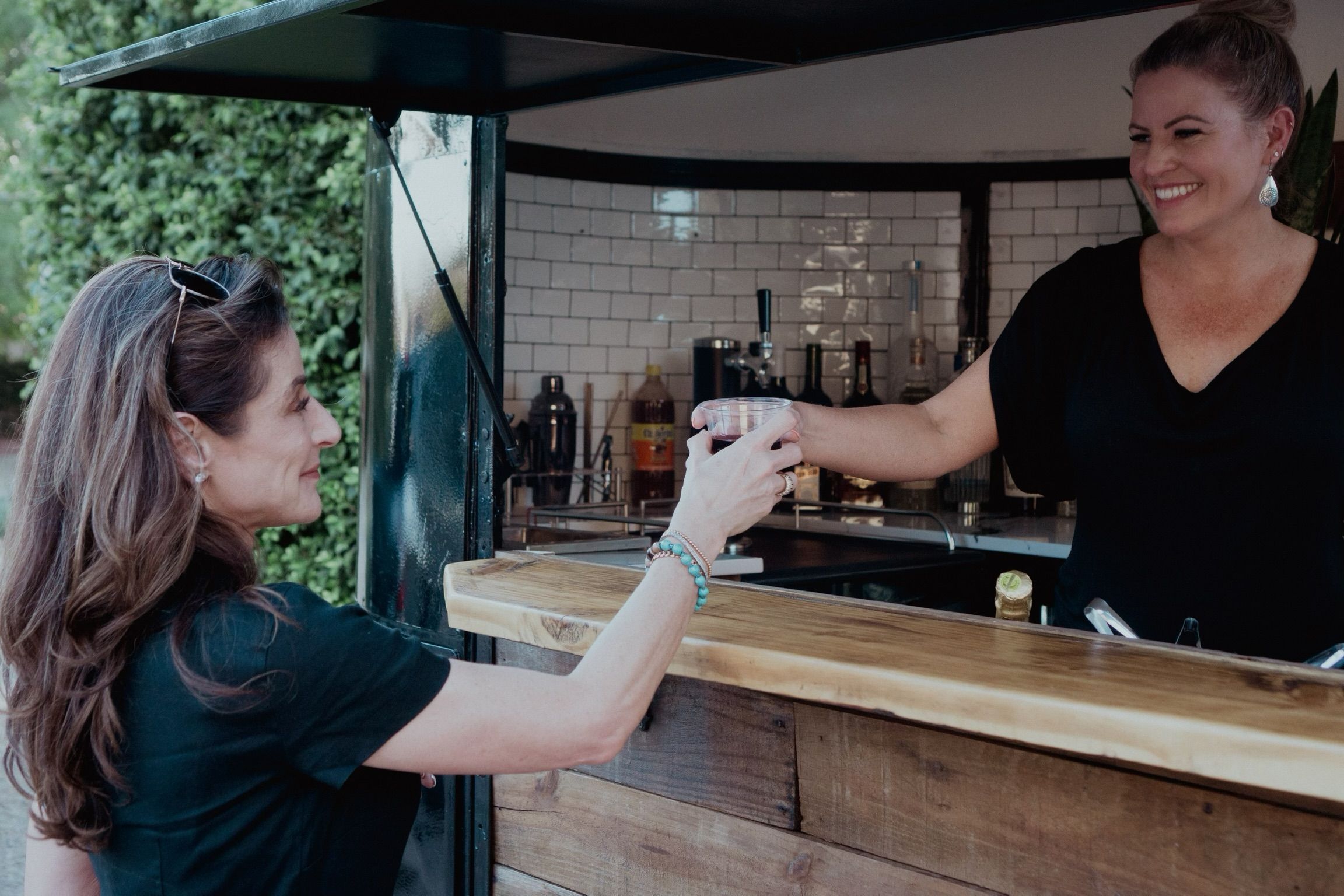 Smiling bartender hands a cold drink to a customer over a rustic wooden outdoor bar with white subway-tile backsplash and greenery in the background.