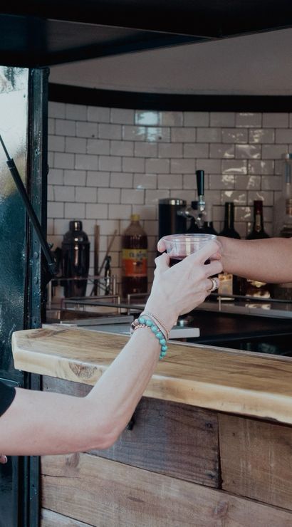 Smiling bartender hands a cold drink to a customer over a rustic wooden outdoor bar with white subway-tile backsplash and greenery in the background.