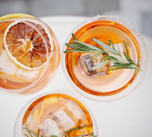 Top-down view of three amber cocktails in clear glasses, garnished with ice cubes, rosemary sprigs and a dried citrus wheel on a bright white surface.