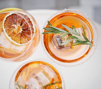 Top-down view of three amber cocktails in clear glasses, garnished with ice cubes, rosemary sprigs and a dried citrus wheel on a bright white surface.