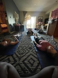 Home yoga and meditation session with participants lying on mats in a cozy apartment living room, white balloon arch by the sliding balcony door, soft daylight and patterned carpet.