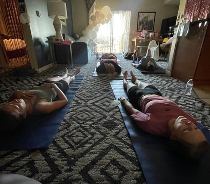 Home yoga and meditation session with participants lying on mats in a cozy apartment living room, white balloon arch by the sliding balcony door, soft daylight and patterned carpet.