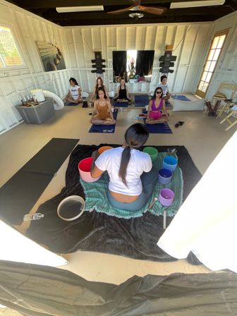 Group yoga meditation class in a bright wooden studio, participants seated on mats for a singing-bowl sound bath while an instructor plays colorful crystal bowls.