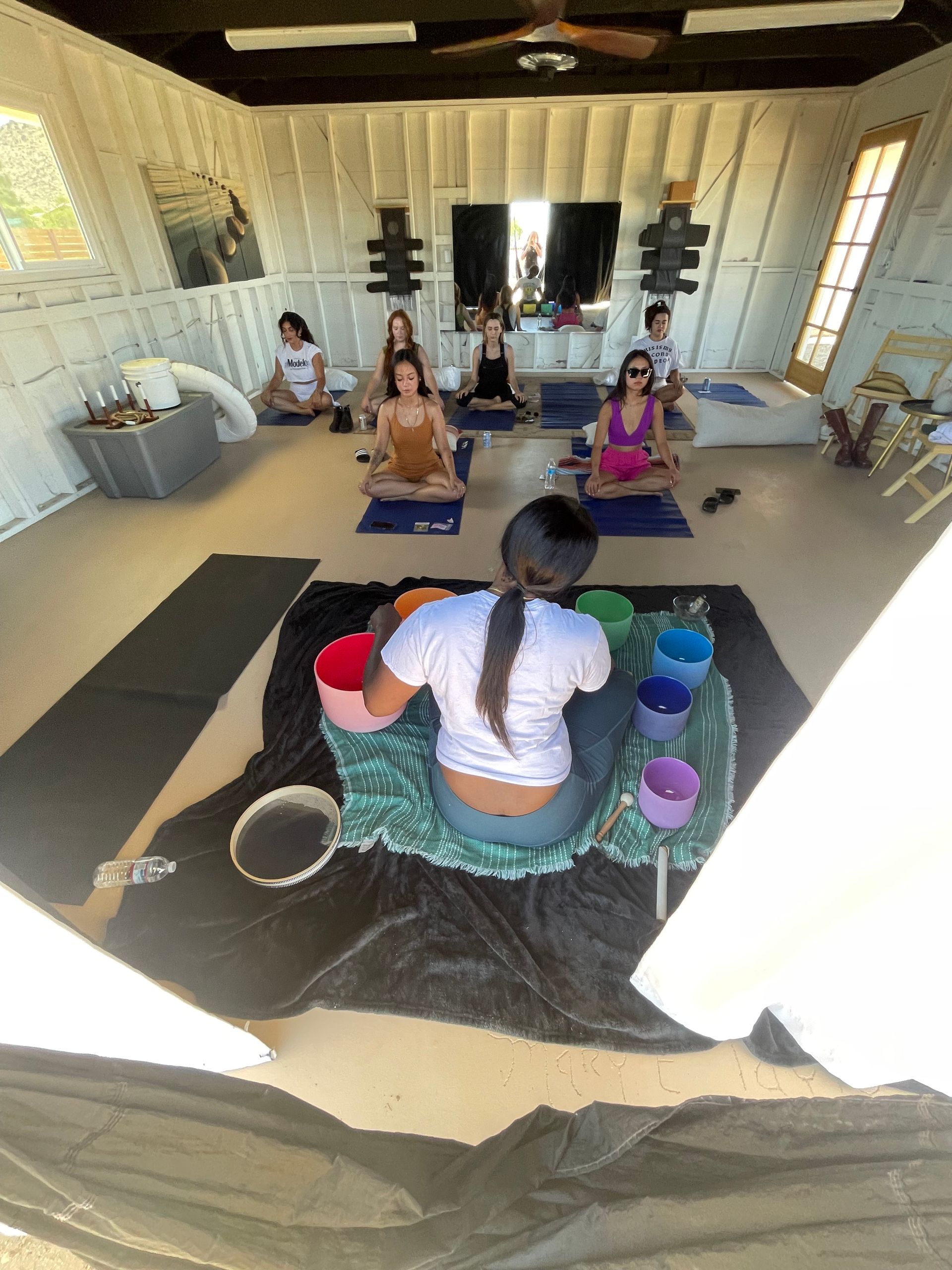 Group sound-bath meditation and yoga class in a bright white-walled studio retreat — instructor with colorful singing bowls in the foreground and participants seated cross-legged on yoga mats.