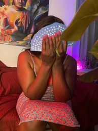 Playful indoor bedroom portrait of a person sitting on a bed, holding a fan of blue star-patterned cards to cover their face; wearing a floral sundress with bright pink nails, colorful Buddha wall art, potted plant and purple ambient lamp in the background.