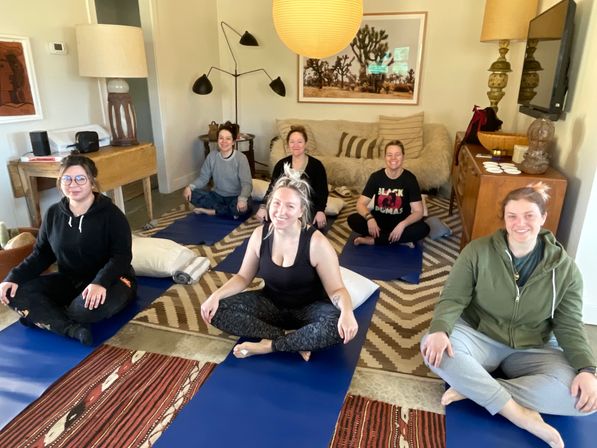 Cheerful home yoga session with six participants seated cross-legged on blue mats in a cozy living room, smiling amid pillows, patterned rugs, wooden furniture and warm pendant lighting.