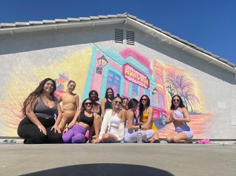Group of women in colorful activewear sitting and kneeling on a sunny pavement in front of a pastel mural on a building wall featuring a retro car, neon-style storefront and palm trees under a bright blue sky.