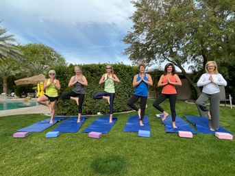 Six women doing tree pose on blue yoga mats on a sunny poolside lawn, colorful activewear, palm trees, hedges and yoga blocks creating a relaxed backyard yoga scene.